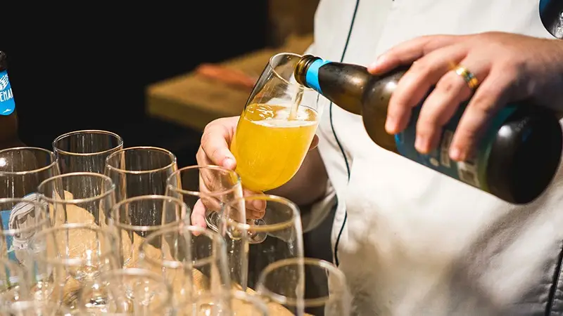 A person, wearing a white shirt and a gold ring, is pouring a yellow beverage from a bottle into a glass. Multiple empty glasses line up on the table in front of them. Among the Best Things to do South Bay October, savoring this moment stands out vividly.