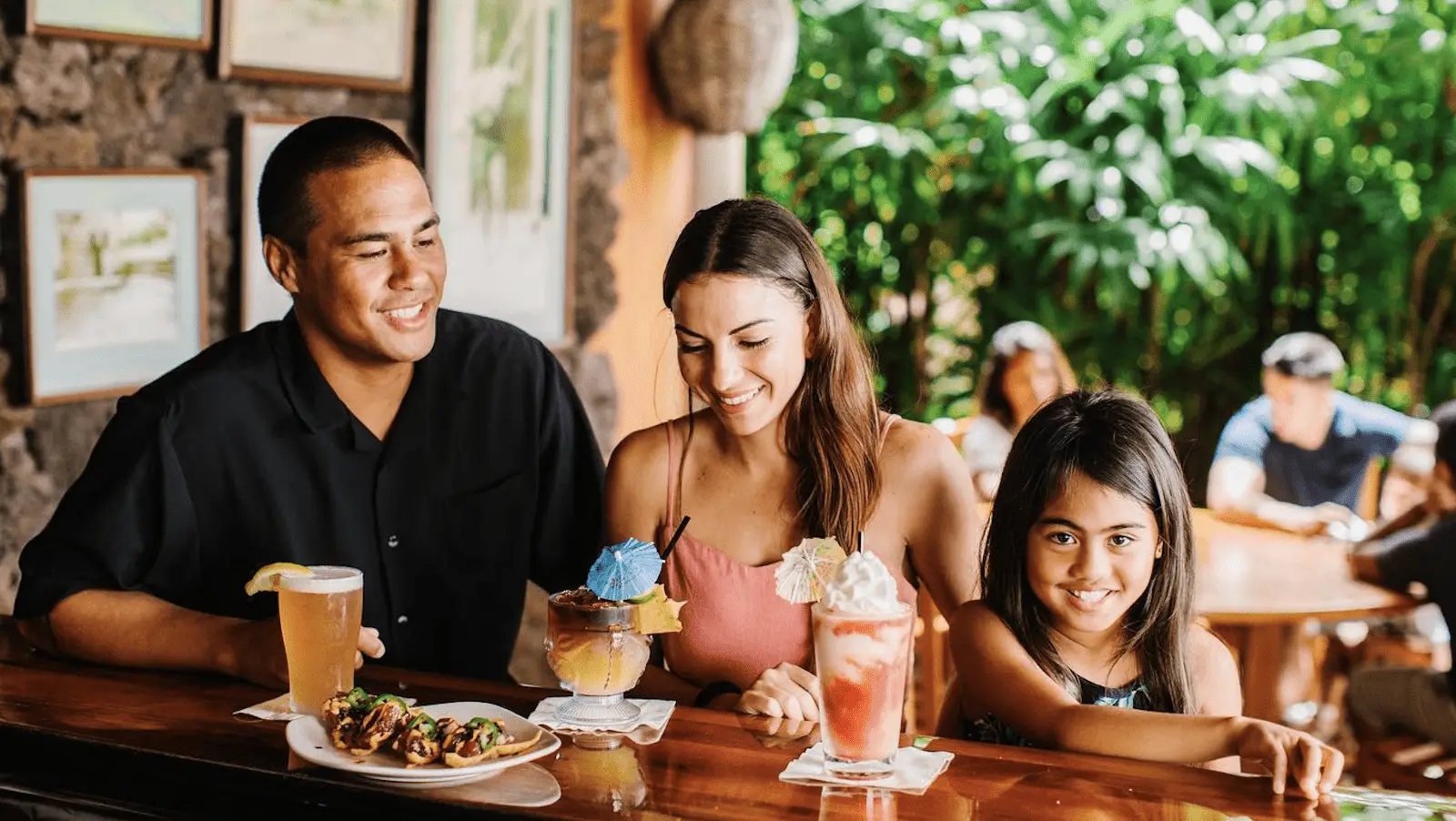 A family of three enjoys a sunset dinner at a tropical-themed restaurant in Kauai. The man has a beer, the woman sips a colorful cocktail with a blue umbrella, and the young girl indulges in a milkshake. Lush greenery and other diners are visible in the background.