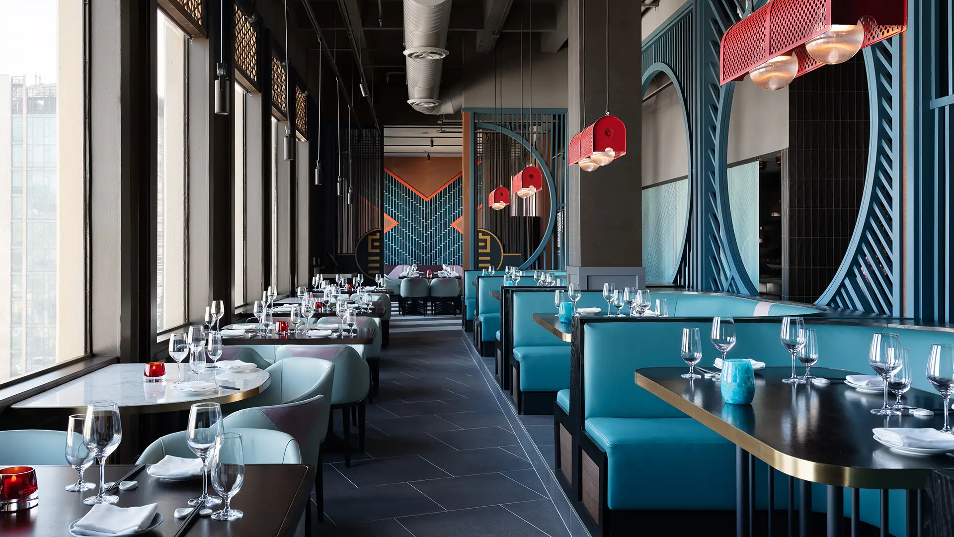 A modern dining area with teal cushioned booths and dark wooden tables set with white plates, wine glasses, and silverware. Red overhead lights hang above the booths. The decor features geometric patterns, large windows, and metal framing with circular cutouts. For the best dinner in San Francisco, look no further.