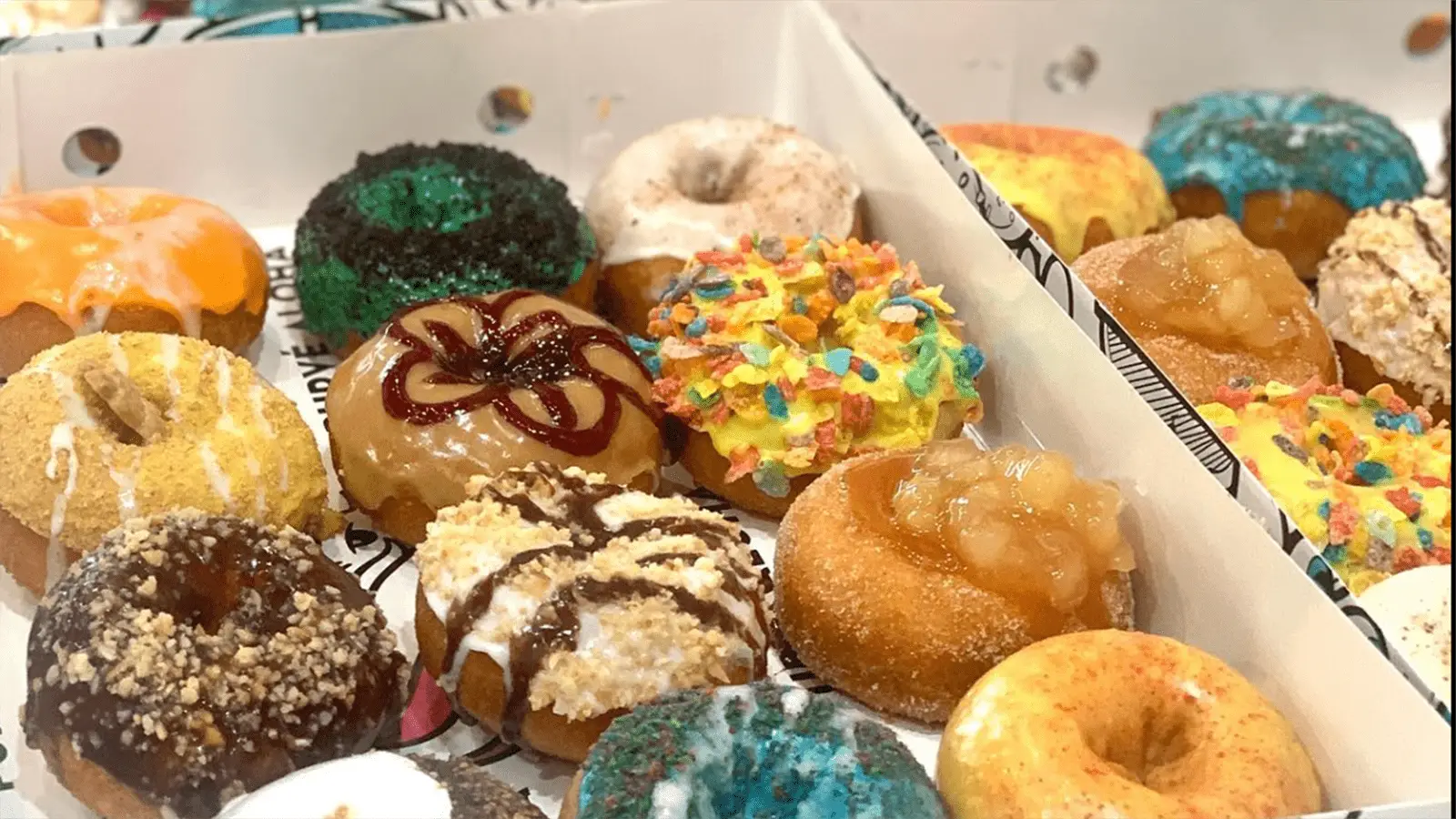 A close-up view of a dozen colorful and assorted donuts in an open white box. The donuts are topped with various glazes, sprinkles, crumbles, and frosting, featuring vibrant colors like blue, yellow, orange, and more. It's no wonder these treats are considered some of the best desserts in Honolulu.