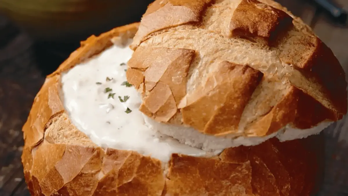 A round loaf of bread with the top cut off, hollowed out, and filled with creamy, white clam chowder reminiscent of the best found on the Monterey Peninsula. The golden-brown crust cradles the soup, which is garnished with a small amount of green herbs, giving a rustic, homemade appearance.