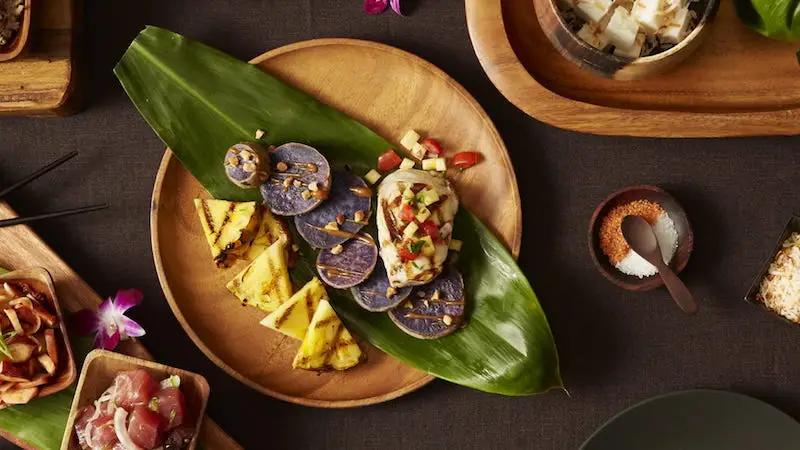 A wooden plate set on a green leaf holds slices of purple sweet potatoes topped with salsa, and wedges of grilled pineapple. Surrounding dishes include bowls with various ingredients and utensils. Purple flowers are scattered throughout the table setting, creating an exotic dining atmosphere reminiscent of where to stay in Wailes.