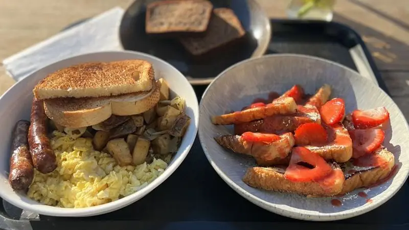 A breakfast tray with two plates: The left plate has scrambled eggs, sausages, diced potatoes, and toast. The right plate has French toast topped with sliced strawberries and syrup, with extra toast in the background—perhaps the best lunch in East Bay for late risers.