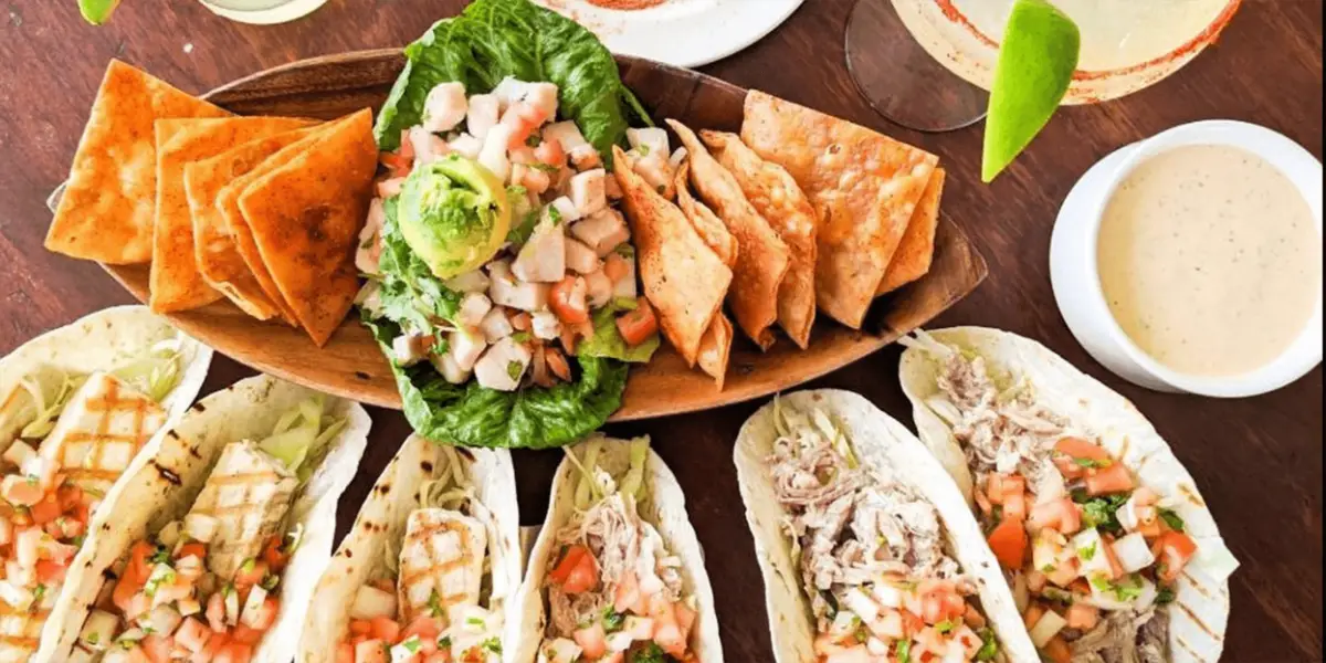 A table spread with various types of tacos filled with grilled chicken and fresh vegetables, accompanied by a platter of tortilla chips, guacamole, and salsa. A drink with a lime wedge and dipping sauce are also visible on the wooden table—perfect for the best sunset dining in Oahu.