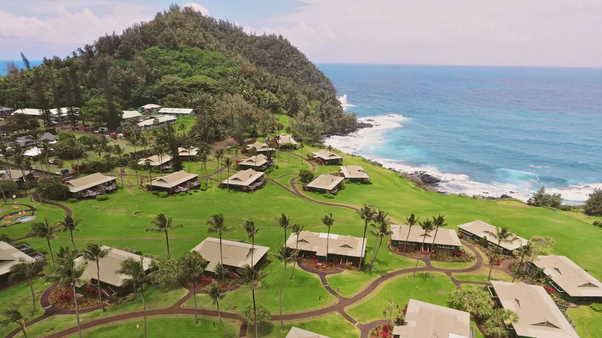 Aerial view of a coastal resort with multiple bungalows surrounded by lush greenery. The resort is located near rocky ocean cliffs with waves crashing against them. There's a hill covered in dense vegetation on one side and clear blue skies above, perfect for mindful travel to Maui.