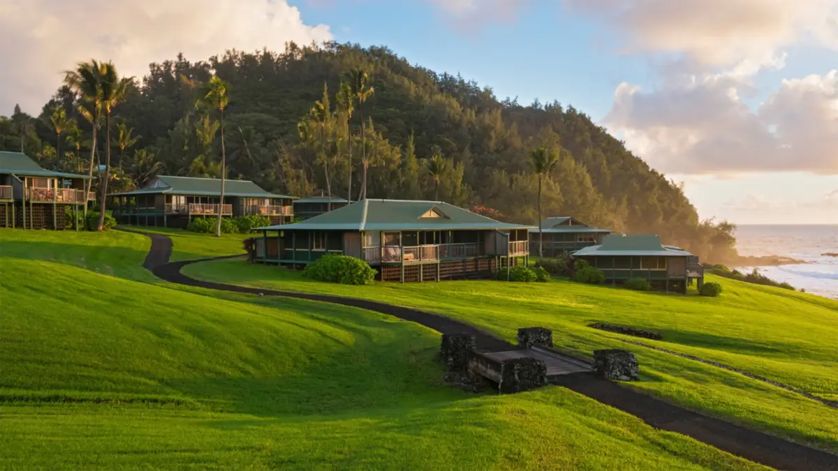 Scenic view of a tropical resort with several green-roofed buildings nestled against a lush hillside. The grounds feature well-maintained lawns, palm trees, and a small bridge over a walkway. For ideas on what to do in Hana, the ocean is visible in the background under a partly cloudy sky.