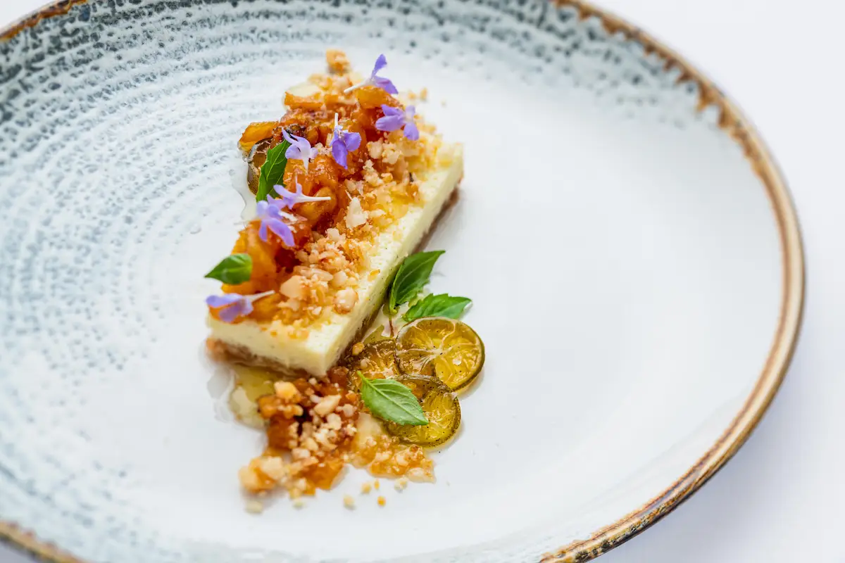 A beautifully plated dish, reminiscent of the Hawaii Food and Wine Festival, features a rectangular slice of creamy dessert garnished with crumbled toppings, small purple flowers, and fresh green herbs. Accompanied by thin slices of citrus and a light syrup, it's presented on a textured white plate with a brown rim.