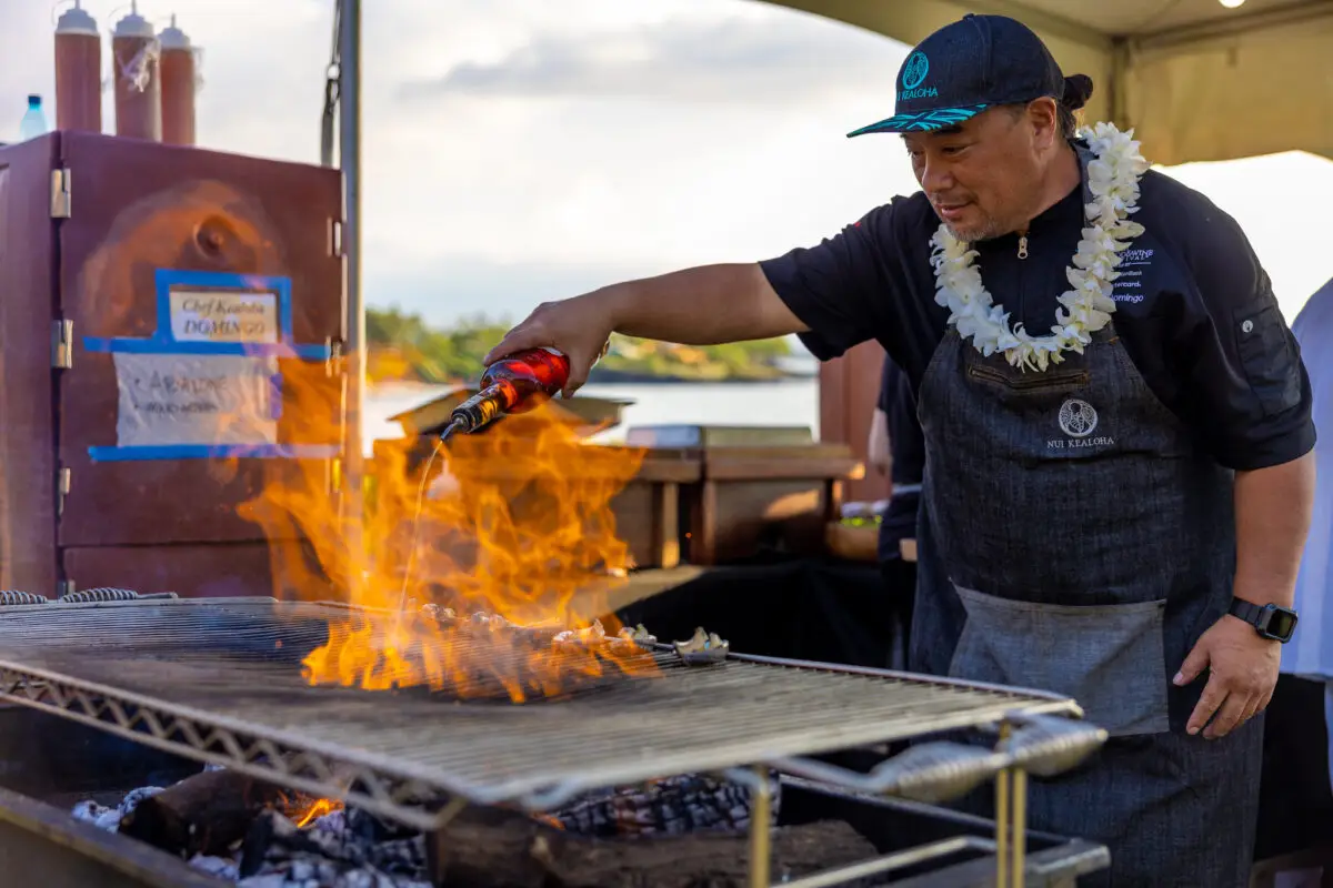 At the Hawaii Food and Wine Festival, a man wearing a hat, an apron, and a lei uses a torch to ignite flames on a grill at an outdoor cooking event. In the background, there is a smoker and other cooking equipment under a canopy. The scene is set against a bright, partially cloudy sky.