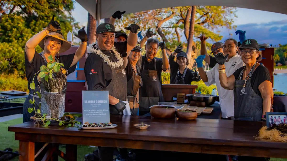 A group of people in chef uniforms and aprons stand together under a tent at the Hawaii Food and Wine Festival, smiling and lifting one arm in celebration. A decorated wooden table in front of them holds various bowls and plates. Trees and a lake are visible in the background.