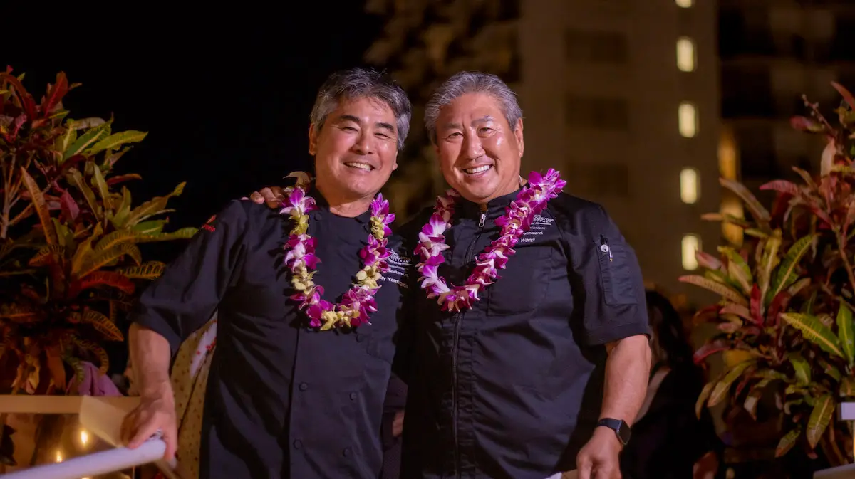 Two smiling men standing side by side, wearing black chef coats and flower leis around their necks. They are outdoors, with greenery and a building lit up in the background. The scene suggests they might be participating in or attending the Hawaii Food and Wine Festival.