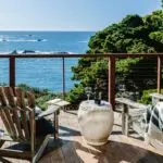 A scenic ocean view from a wooden deck with two Adirondack chairs facing the water. A small round table between the chairs holds two wine glasses and a bottle of wine. A soft blanket is draped over one chair, and lush green trees frame the background—an ideal spot if you're wondering where to stay in North Bay.