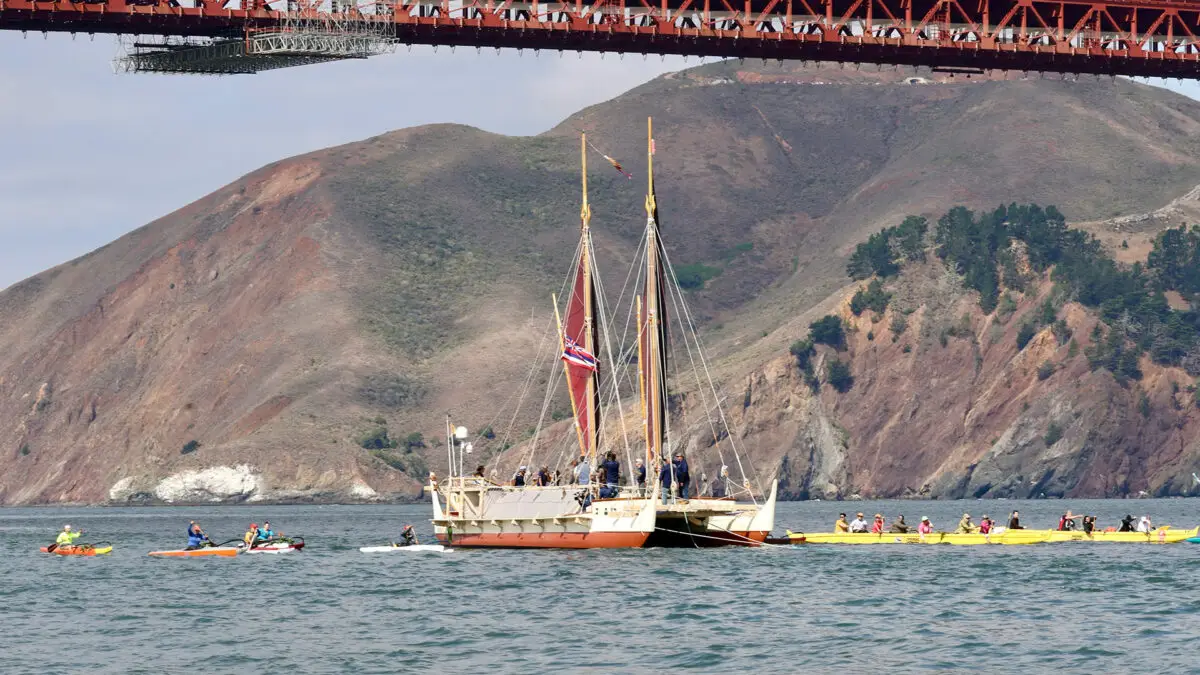 A large sailboat with red sails, resembling the Hokule'a, is flanked by several kayaks and smaller boats, navigating under a red-brick bridge against a backdrop of green and brown hills in Sausalito Marin. The sky is partly cloudy.