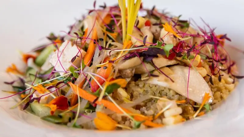 A colorful quinoa salad topped with sprouts, microgreens, sliced vegetables, and edible flowers. The dish is artistically arranged on a white plate, showcasing a vibrant and fresh presentation—perfect for a Sunset Dinner in Kauai.