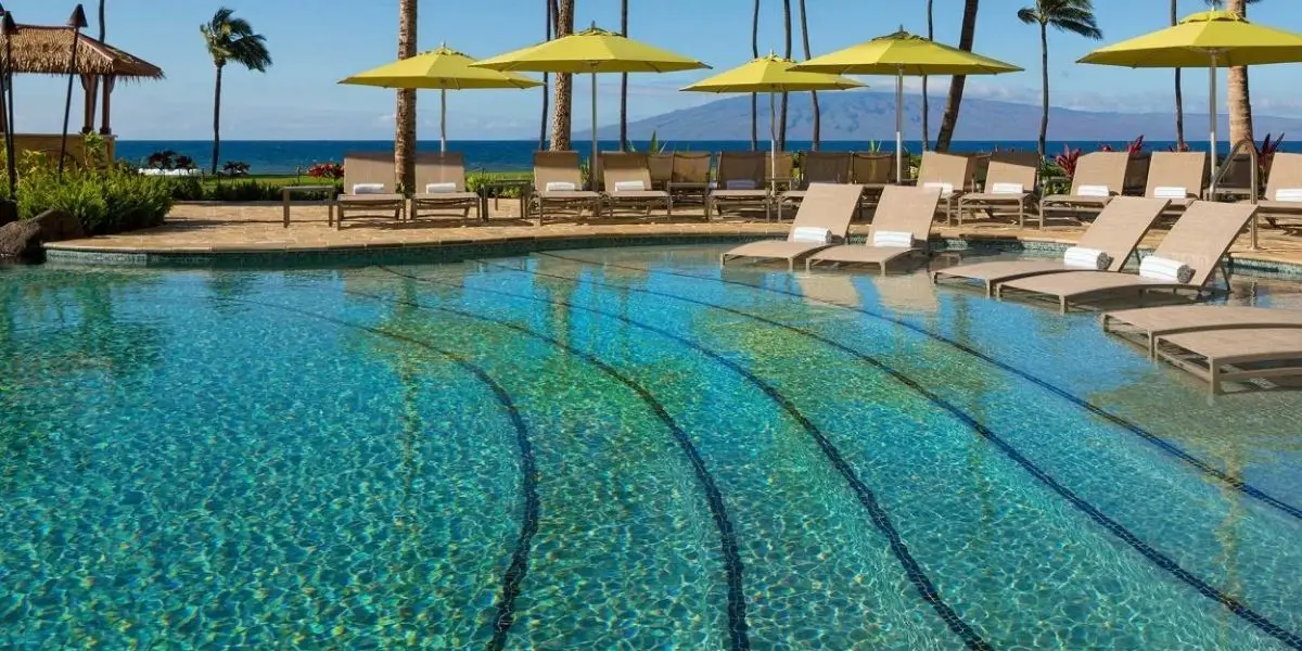 A pristine outdoor pool at the Hyatt Residences has multiple lounge chairs and yellow umbrellas on a sunny day. The pool features wide, shallow entry steps. Palm trees, a grass hut, and the ocean are visible in the background, with a clear blue sky overhead.