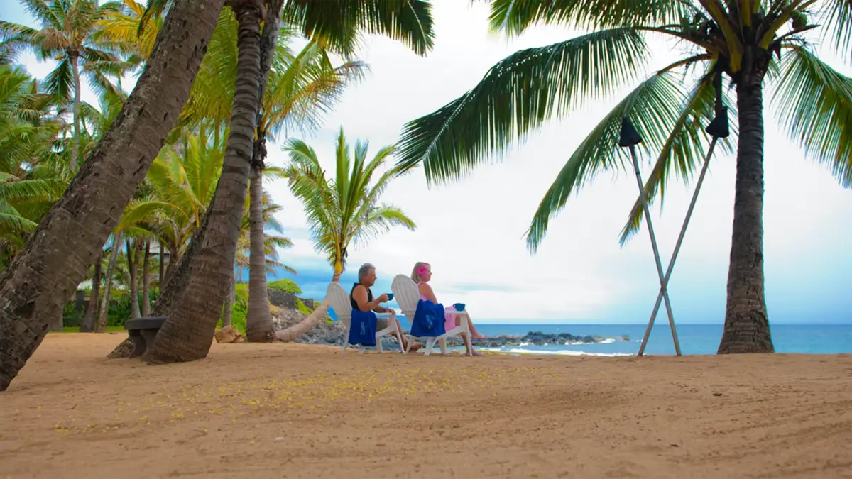 Two people sit on beach chairs under palm trees, facing the ocean, at one of the best romantic hotels in Maui. They are enjoying a calm, sandy beach with gentle waves in the background. The sky is bright with some clouds. The scene is serene and tropical, suggesting a relaxing vacation spot.