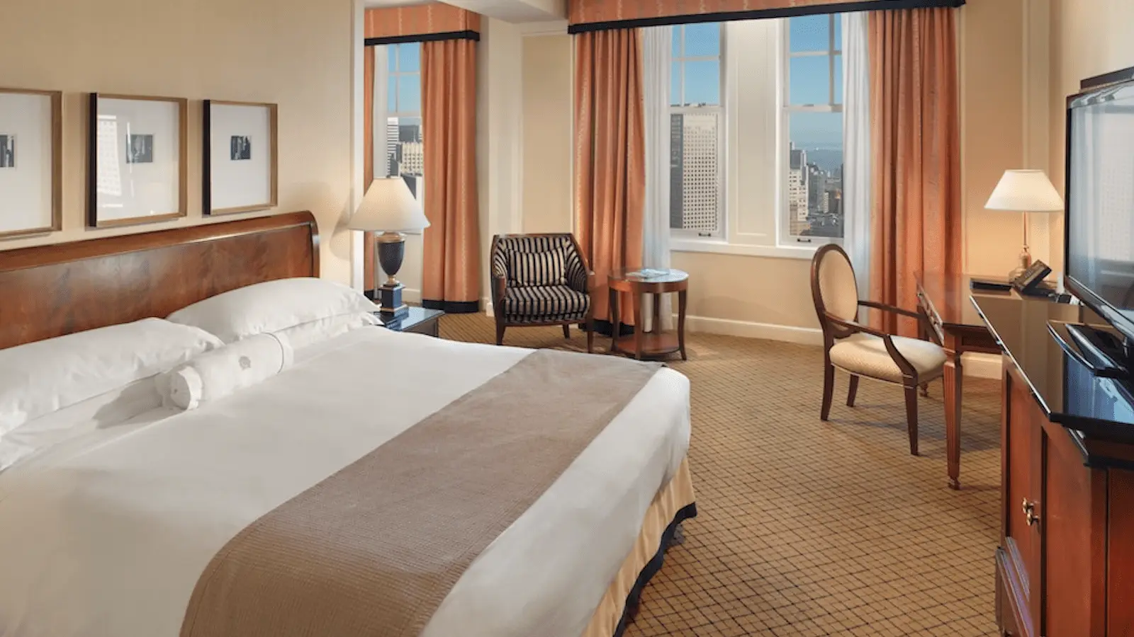 A spacious hotel room featuring a large bed with white linens and a beige throw. The room has a window with city views, peach-colored curtains, framed wall art above the bed, a black and white striped armchair, a round table, and a flat-screen TV on a stand. Perfect for those seeking the Best Spa and Wellness Hotels in the Bay Area.