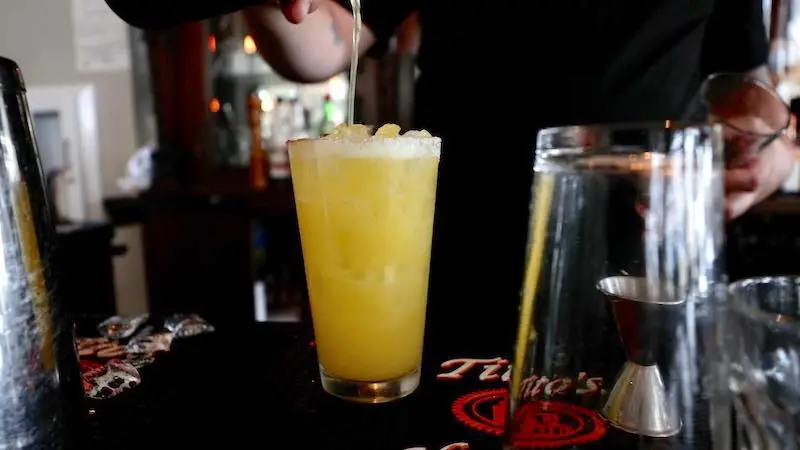 A bartender is preparing a cocktail during the best happy hour on the Big Island. They are pouring a liquid into a tall glass filled with ice and a yellow beverage, possibly orange juice. Various bartending tools and glassware are placed on the counter.