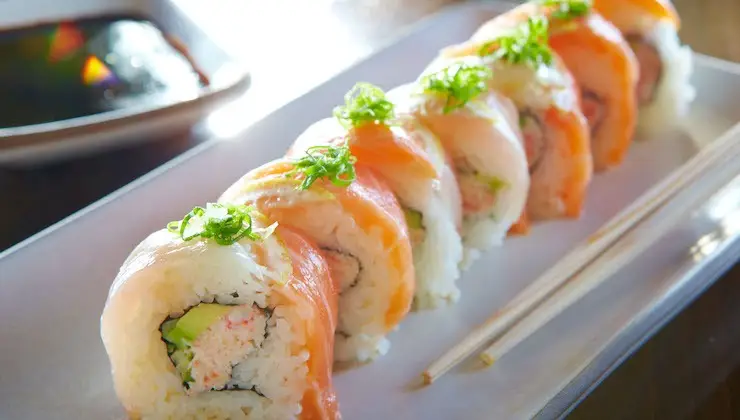 A close-up of a sushi roll arranged on a white plate, garnished with green onion. The roll includes pieces of fish, avocado, and rice, reminiscent of the flavors at Best Poke Maui. A side dish of soy sauce and a pair of wooden chopsticks are placed beside the plate.