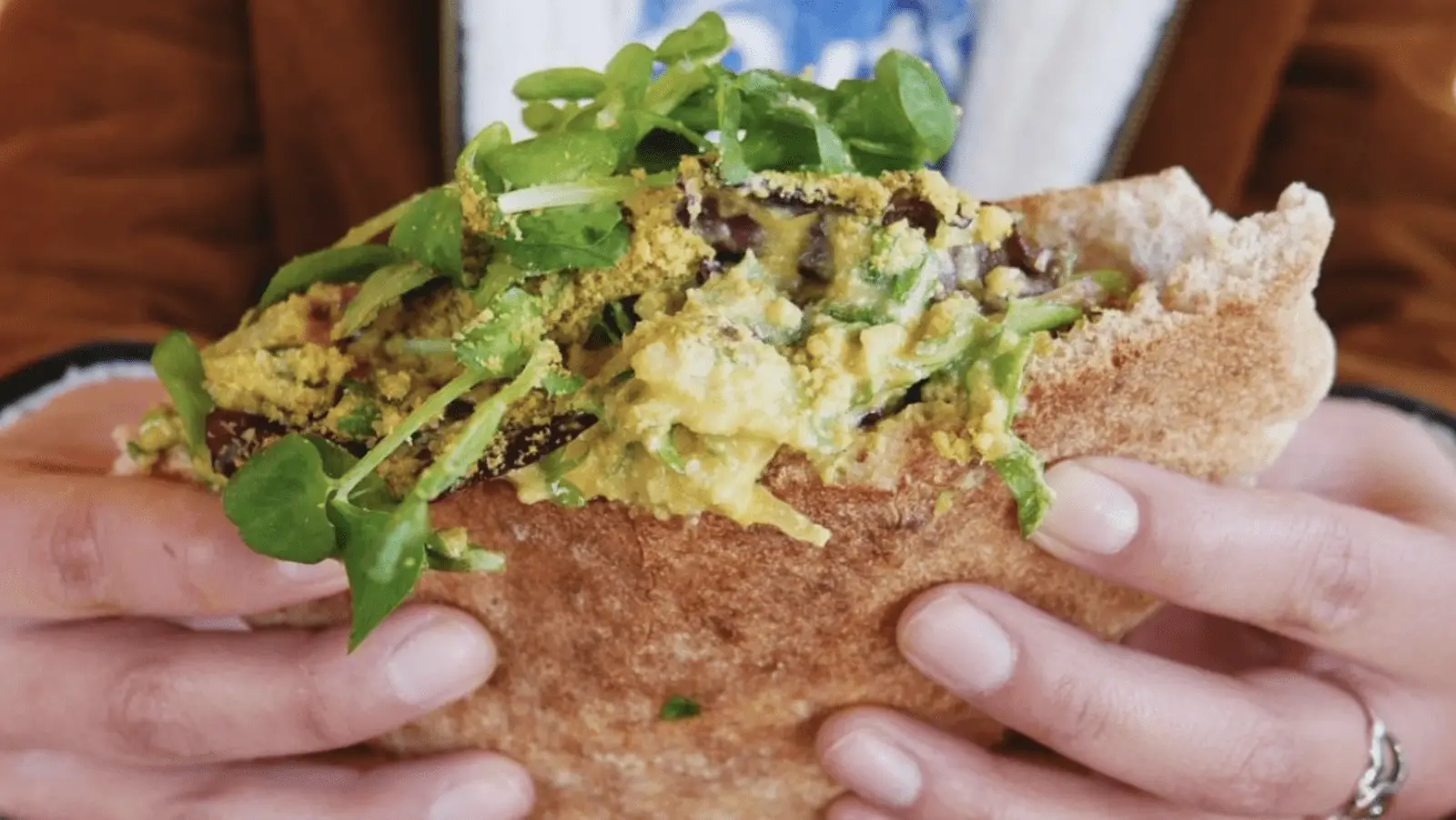 Close-up of a person holding a pita bread sandwich filled with greens and a mixture of ingredients, including what appears to be avocado and other chopped vegetables. The person’s hands are visible, and they are wearing a brown jacket and white shirt.