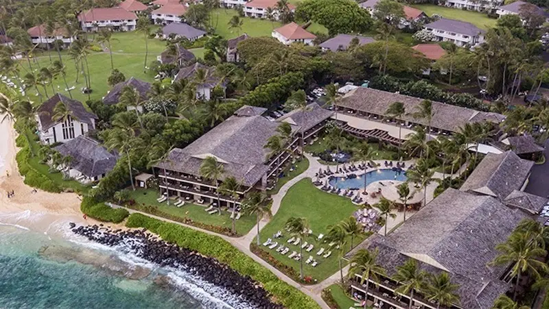 Aerial view of a tropical resort by the beach with lush greenery, several buildings, a pool area, and lounge chairs arranged on the grass and around the pool. The shoreline is sandy with rocks near the water, and waves can be seen gently crashing—a perfect example of hotels with spas in Hawaii.