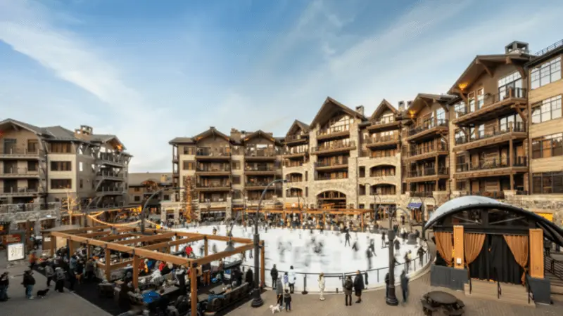 A bustling outdoor ice skating rink is surrounded by timber-framed buildings under a blue sky. Skaters fill the rink while spectators watch from nearby. The buildings have a rustic, chalet-style design, contributing to a cozy and inviting atmosphere, making it one of the best things to do in Tahoe January.