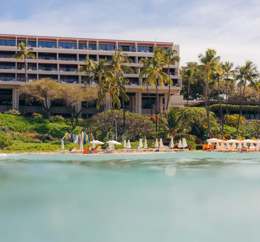 View of the best oceanfront hotel on Kohala Coast, surrounded by lush greenery and tall palm trees. The hotel has several stories with balconies. In the foreground are beach chairs and umbrellas along the sandy shore, and the clear blue water of the ocean is visible.