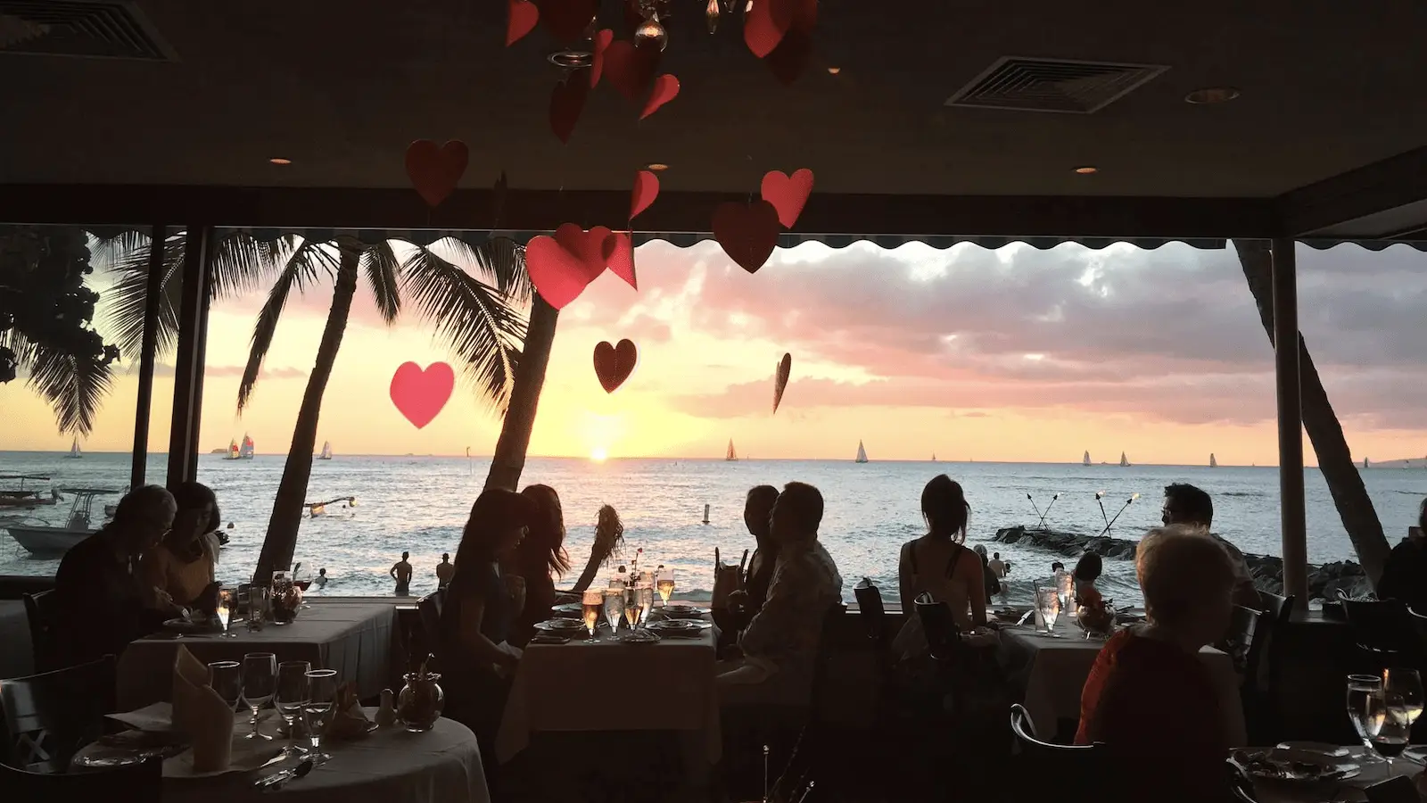 People dining at a restaurant with large windows overlooking a stunning sunset view of the ocean. Silhouettes of sailboats can be seen on the horizon. Red heart decorations hang from the ceiling, creating a romantic atmosphere. Palm trees are visible outside, making it the best spot for sunset dining in Oahu.