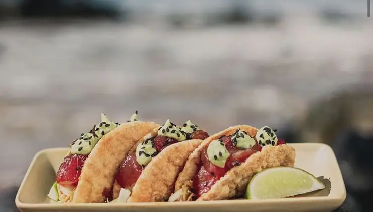 A plate of three tacos filled with raw fish, diced cucumbers, and garnished with sesame seeds, served with a wedge of lime on the side. The background shows a blurred view of what appears to be a beach in Lahaina. This might just be the best dinner spot near Kapalua or Kanapali.