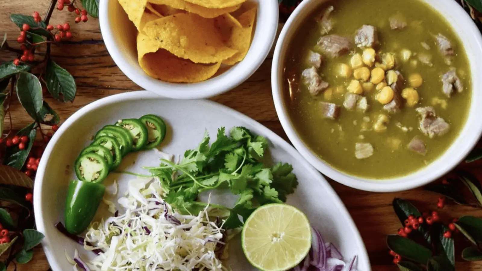 A wooden table features a bowl of green soup with corn and chunks of meat, a bowl of tortilla chips, and an oval plate with sliced jalapeños, cilantro, shredded cabbage, red onion, and a halved lime. Small sprigs with red berries frame the scene—truly the best lunch San Francisco has to offer.