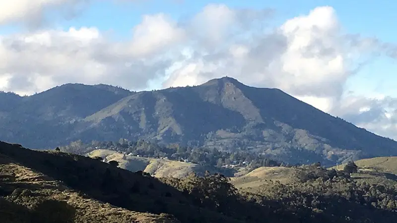 A scenic view of a mountain range with a prominent peak under a partly cloudy sky. The foreground includes rolling hills with patches of greenery and some trees, perfect for outdoor activities. Shadows from scattered clouds create a mottled effect on the hillsides, inviting exploration akin to north bay things to do.