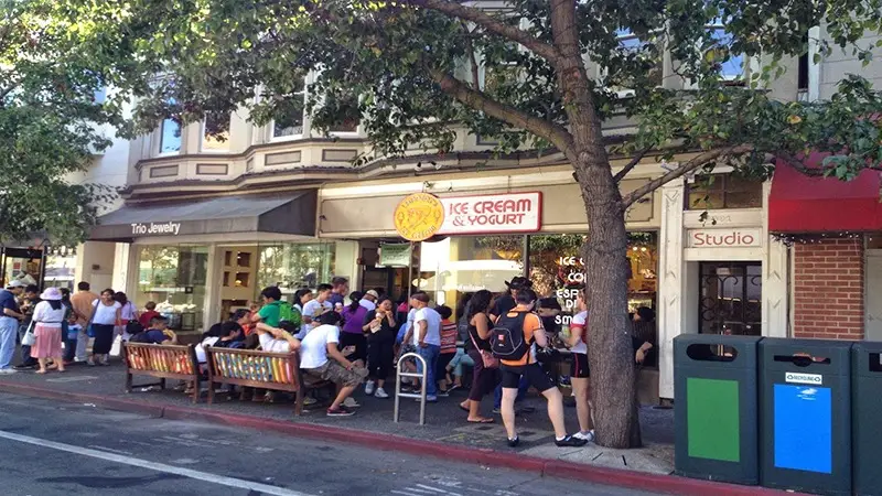 A bustling street scene with a crowd of people standing in line outside an ice cream and yogurt shop. The shop is flanked by jewelry and studio storefronts. Benches and trees line the sidewalk, and there are recycling bins in the foreground—perfect for those looking to explore East Bay activities.