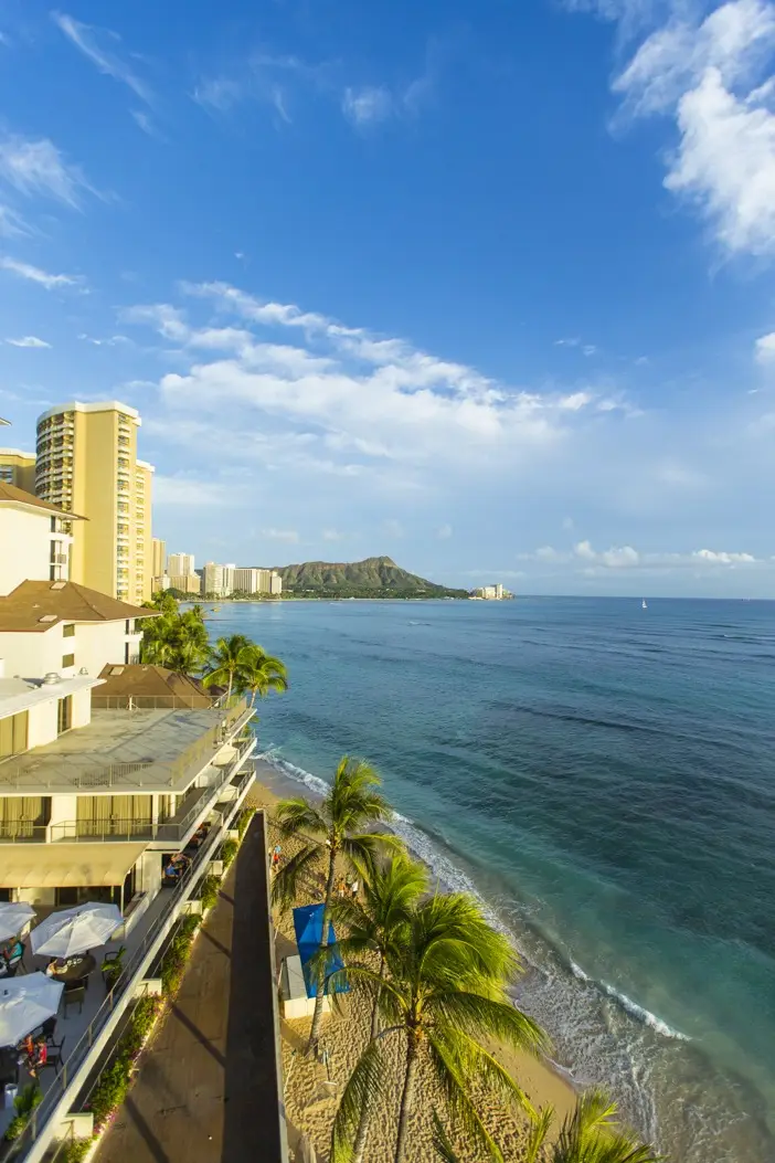 A scenic view of Waikiki Beach, Hawaii, showcasing sandy shores lined with palm trees and lapped by clear blue waters. High-rise buildings are visible on the left, and Diamond Head, an iconic volcanic tuff cone, stands prominently in the background under a partly cloudy sky.