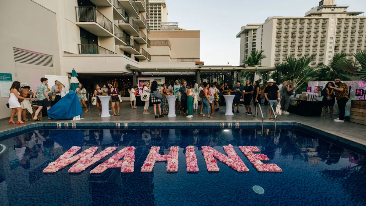 A diverse group of people gathers by a pool at one of the luxurious Outrigger Hotels on Oahu, socializing and enjoying an outdoor event. The pool features a floating decoration with "WAHINE" spelled out in pink flowers, while surrounding buildings and palm trees create a modern, tropical setting.