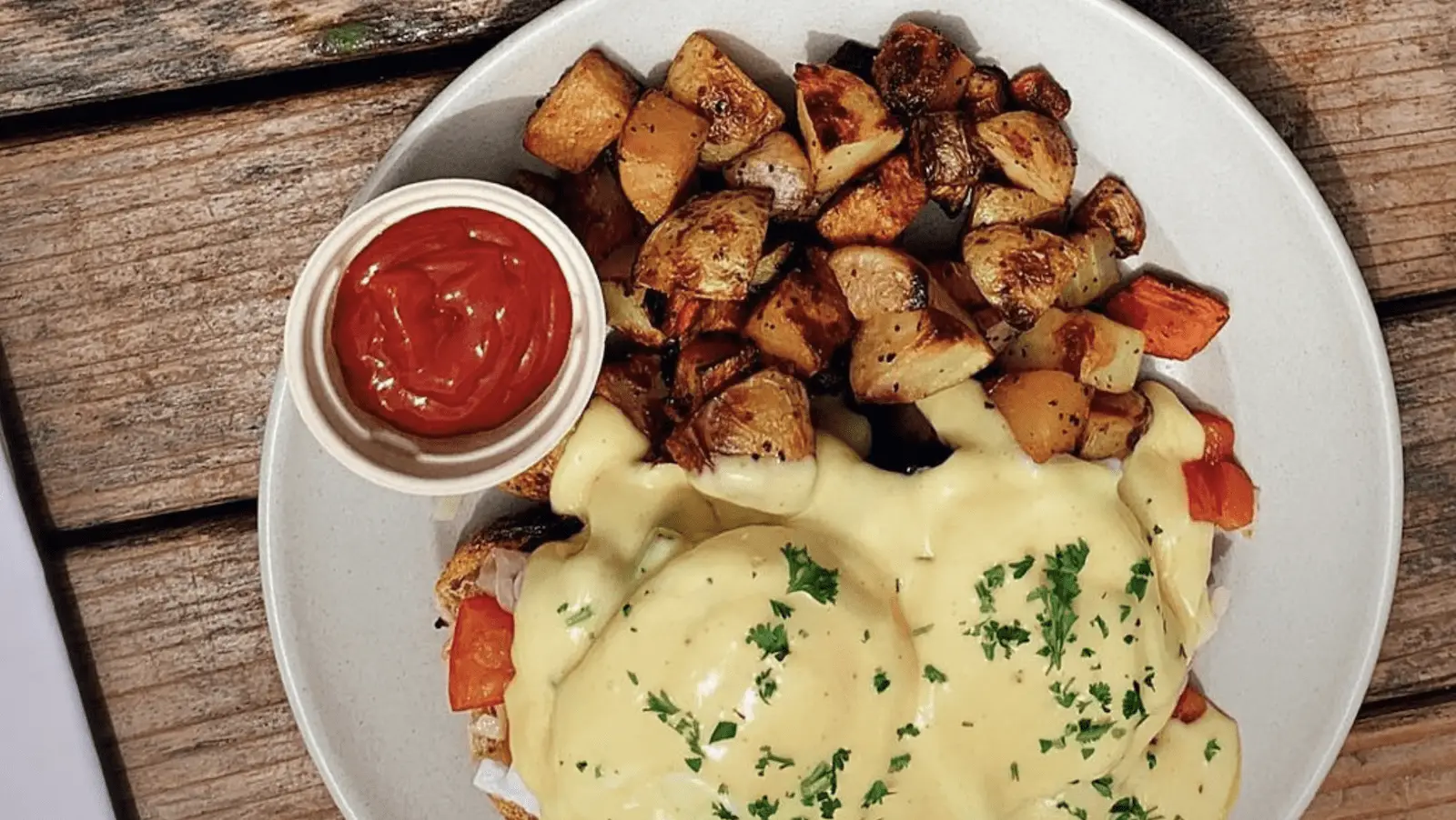 A white plate on a wooden table contains a dish of seasoned roasted potatoes, a poached egg covered in hollandaise sauce, and garnished with chopped parsley. A small container of ketchup is placed on the side—truly the best breakfast North Bay has to offer.