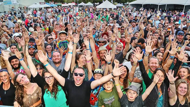 A large crowd of enthusiastic people of various ages is gathered outdoors at one of the Best Annual Events in the North Bay. Many are smiling, waving, and raising their hands towards the camera. Some are wearing colorful clothes and accessories. White tents are visible in the background along with a stage.