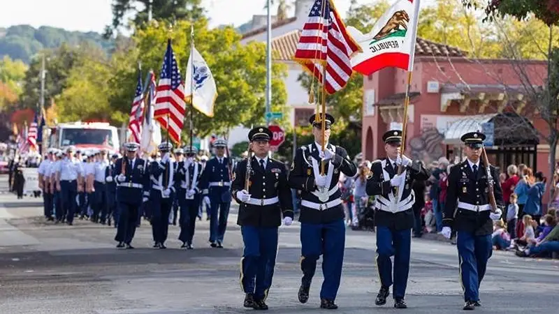 A parade with military personnel marching in uniform, carrying American and Californian flags. Crowds of spectators line the street, watching the procession. Trees and buildings are visible in the background, making it one of the Best Annual Events in the North Bay.