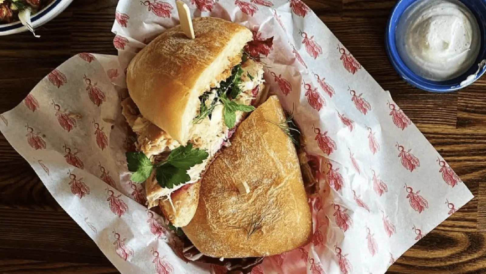 A large sandwich, arguably the best lunch in San Francisco, sits on deli paper printed with red cows, on a wooden table. The sandwich has shredded meat and fresh greens on a crusty roll, with a toothpick holding it together. A small blue bowl of white dipping sauce is placed nearby.