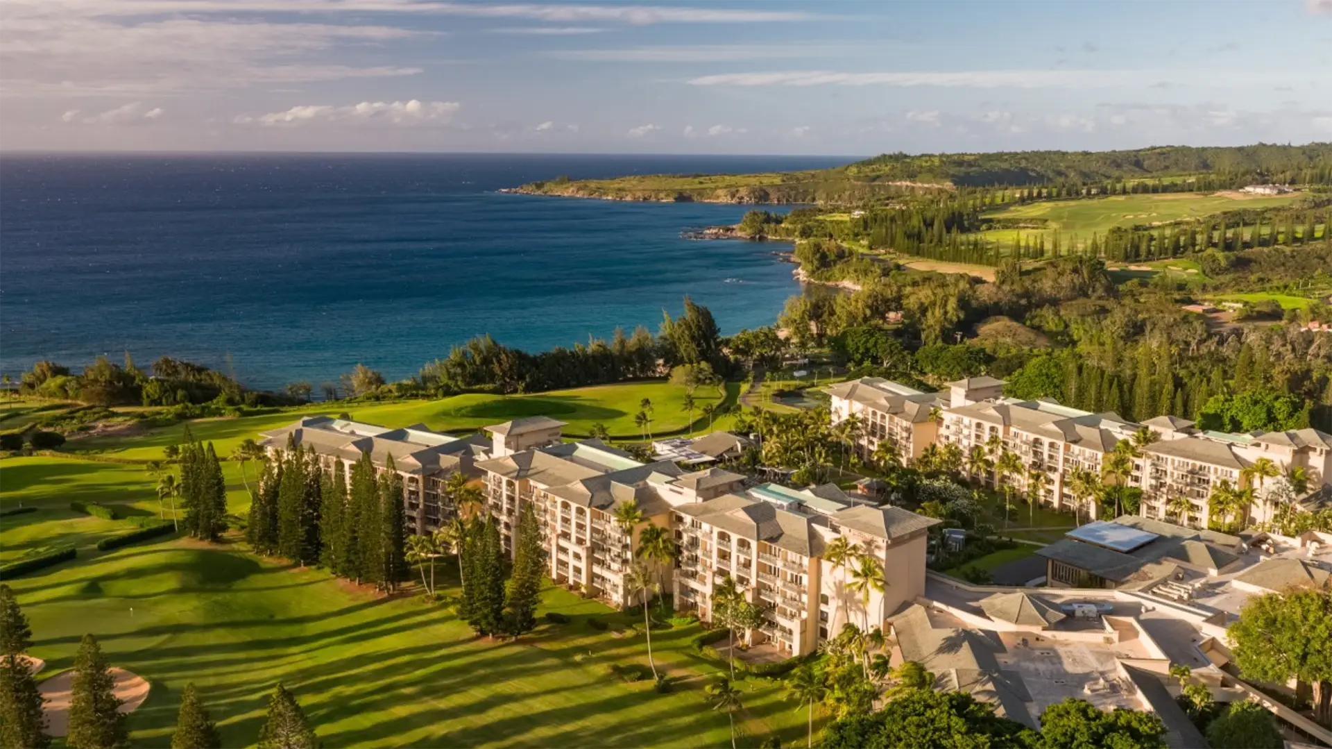 Aerial view of one of the best luxury hotels in West Maui, surrounded by lush greenery and situated on an oceanfront property. Several buildings with white facades and balconies are visible, with manicured lawns, tall trees, and the coastline extending into the horizon under a partly cloudy sky.