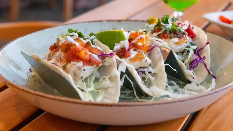 A plate of three tacos filled with fish, topped with shredded cabbage, diced tomatoes, creamy white sauce, and cilantro, garnished with a lime wedge, sits on a wooden table. It's the perfect dish to enjoy during the best happy hour on the Big Island.