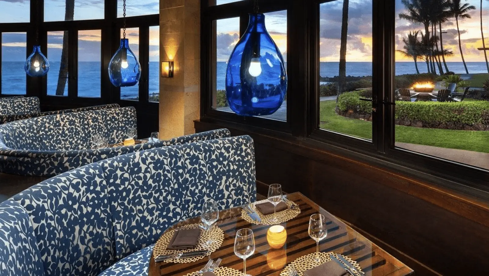 A cozy restaurant with blue, patterned booth seating overlooks a breathtaking coastal view during sunset dinner in Kauai. Large blue pendant lights hang from the ceiling. The tables are set with glasses, plates, and folded napkins. Palm trees are visible outside the window.