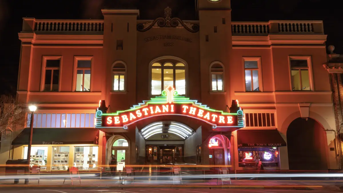 A historic building lit up at night with a neon sign reading "Sebastiani Theatre". The theater's entrance is flanked by two windows glowing from interior lights, and the street in front shows trails of light from passing vehicles. The building, known for hosting some of the best annual events in Sonoma County, has exquisite architectural detailing.