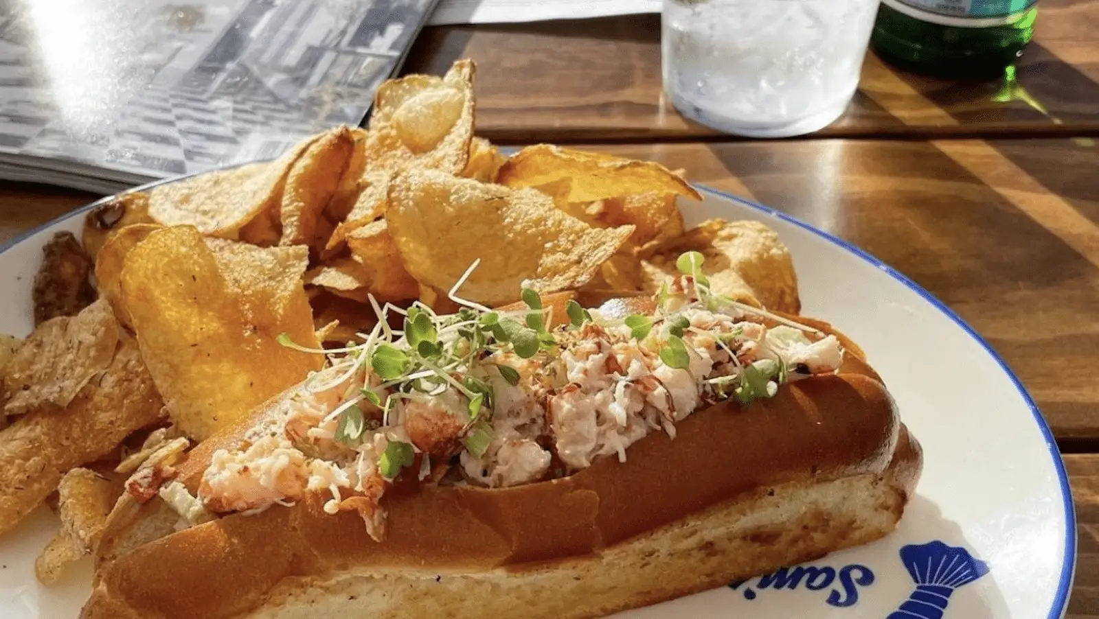 A plate featuring a lobster roll topped with microgreens is served with a side of potato chips. Resting on a wooden table alongside a glass of water, an open magazine, and a green bottle, the meal is bathed in sunlight. It could easily be considered one of the best breakfasts in North Bay.