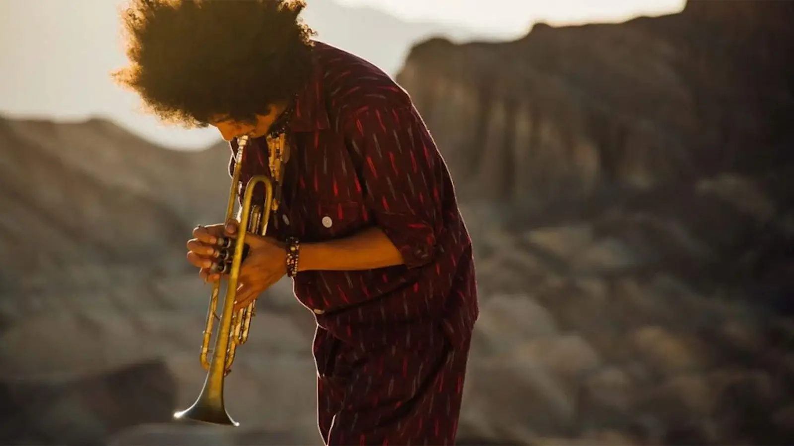 A person with an afro hairstyle plays a trumpet outdoors in a rocky, desert-like landscape during the day. Dressed in a red, patterned outfit and appearing deeply engaged in their music, they showcase one of the best things to do in the South Bay this August.