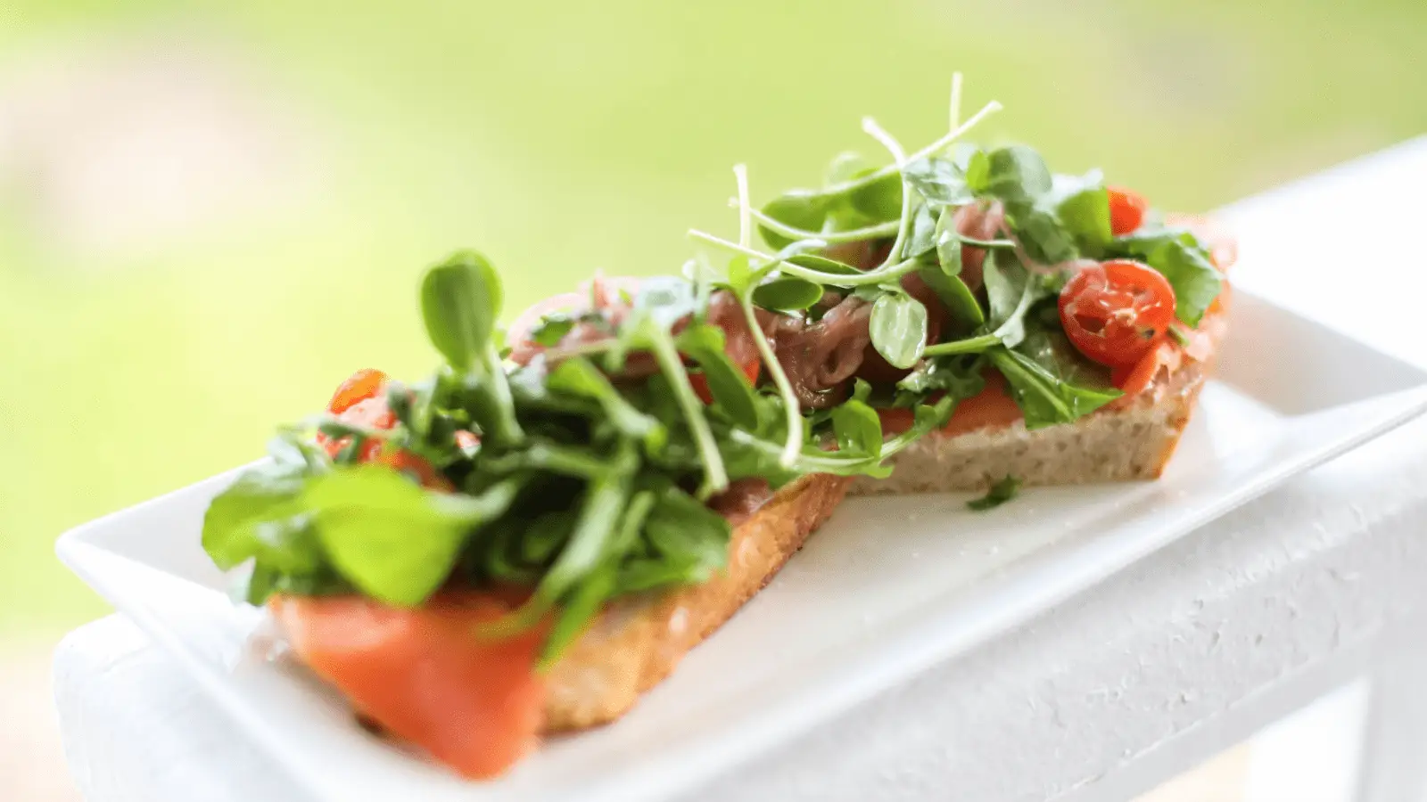 An open-faced sandwich on a white rectangular plate is topped with smoked salmon, cherry tomatoes, microgreens, and arugula. The background is blurred, highlighting the vibrant colors of the fresh ingredients—truly the Best Breakfast Kauai has to offer.