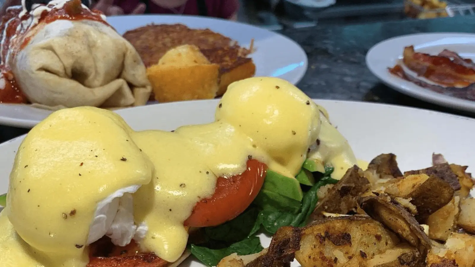 A close-up of a plate with eggs Benedict featuring poached eggs, hollandaise sauce, fresh spinach, and tomato slices, alongside a generous serving of home fries. In the background, there's a plate with a burrito topped with sauce and hash browns—truly the Best Breakfast Kauai has to offer.