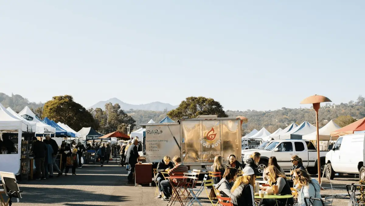 A vibrant outdoor market scene with people sitting at colorful tables, eating, and socializing. Food trucks and stalls line both sides of the walkway amidst a backdrop of mountains and trees under a clear sky—perfect for those looking to explore East Bay activities.