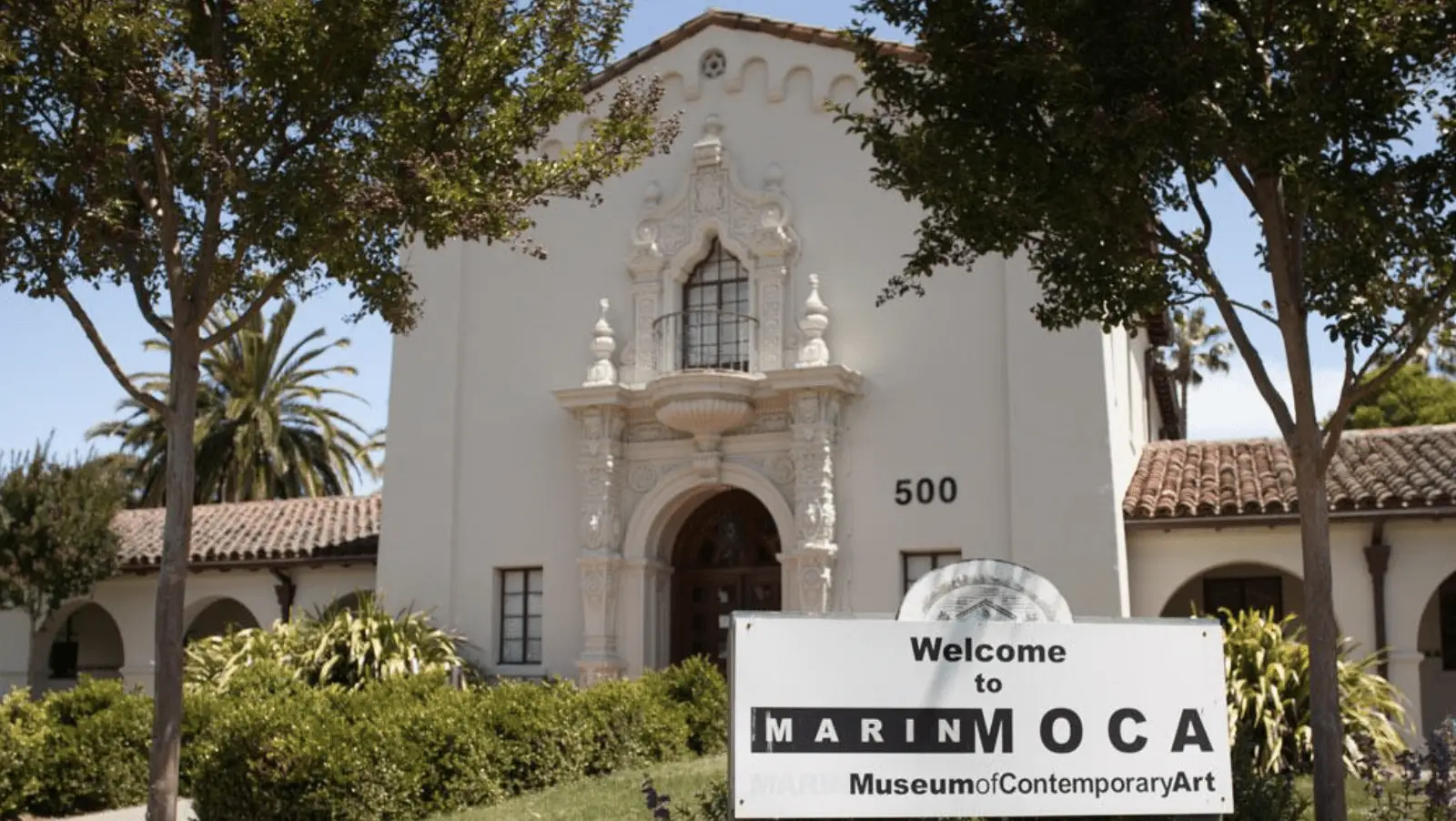 The Marin Museum of Contemporary Art, a white stucco building with ornate architectural details and arched windows, is partially obscured by trees. A sign in front reads "Welcome to Marin MOCA Museum of Contemporary Art" with the number 500 above the entrance. North Bay things to do include visiting this artistic gem.