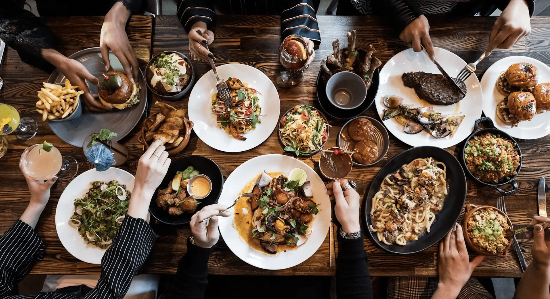 A wooden table is covered with a variety of dishes including salads, burgers, fries, steak, chicken wings, seafood, and cocktails. Several pairs of hands are seen reaching for and sharing the food, evoking a Bay Area Thanksgiving Offering that highlights the joy of communal dining.