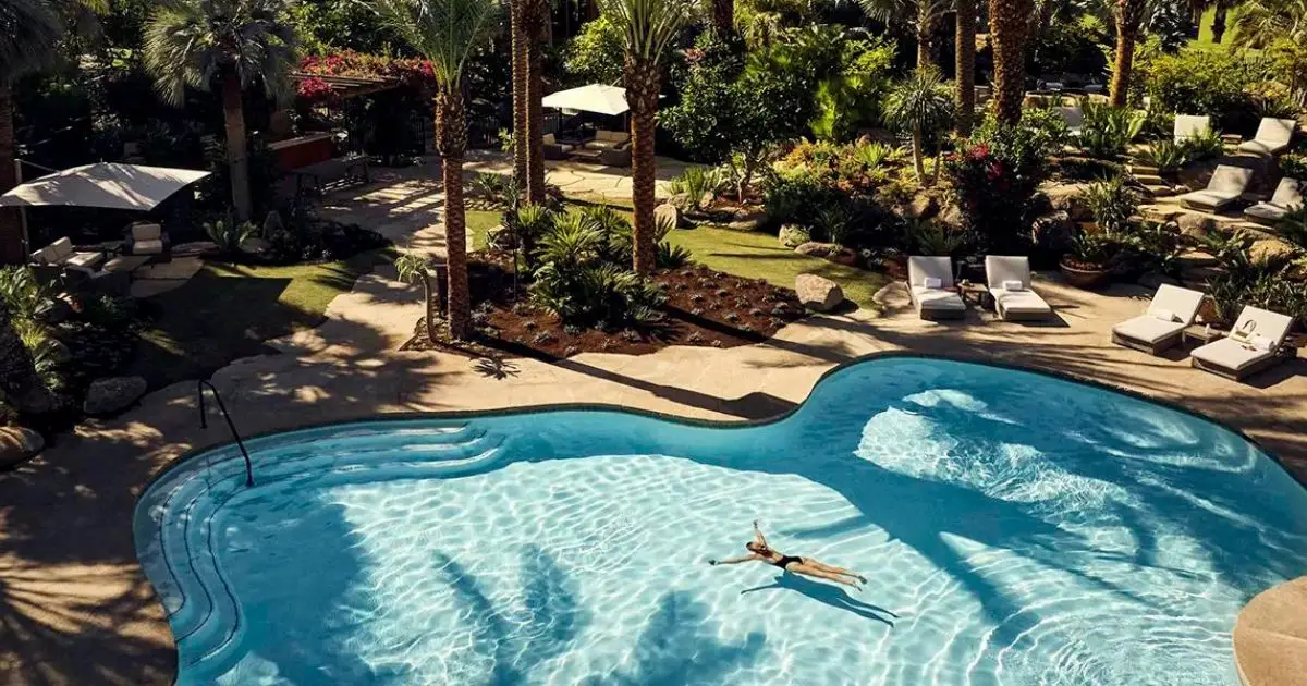 An aerial view of a serene outdoor pool surrounded by lush palm trees and greenery, epitomizing one of the best hotel pools in California. A person leisurely floats on their back in the pool, while several lounge chairs and umbrellas are scattered around the poolside, creating a relaxing atmosphere.