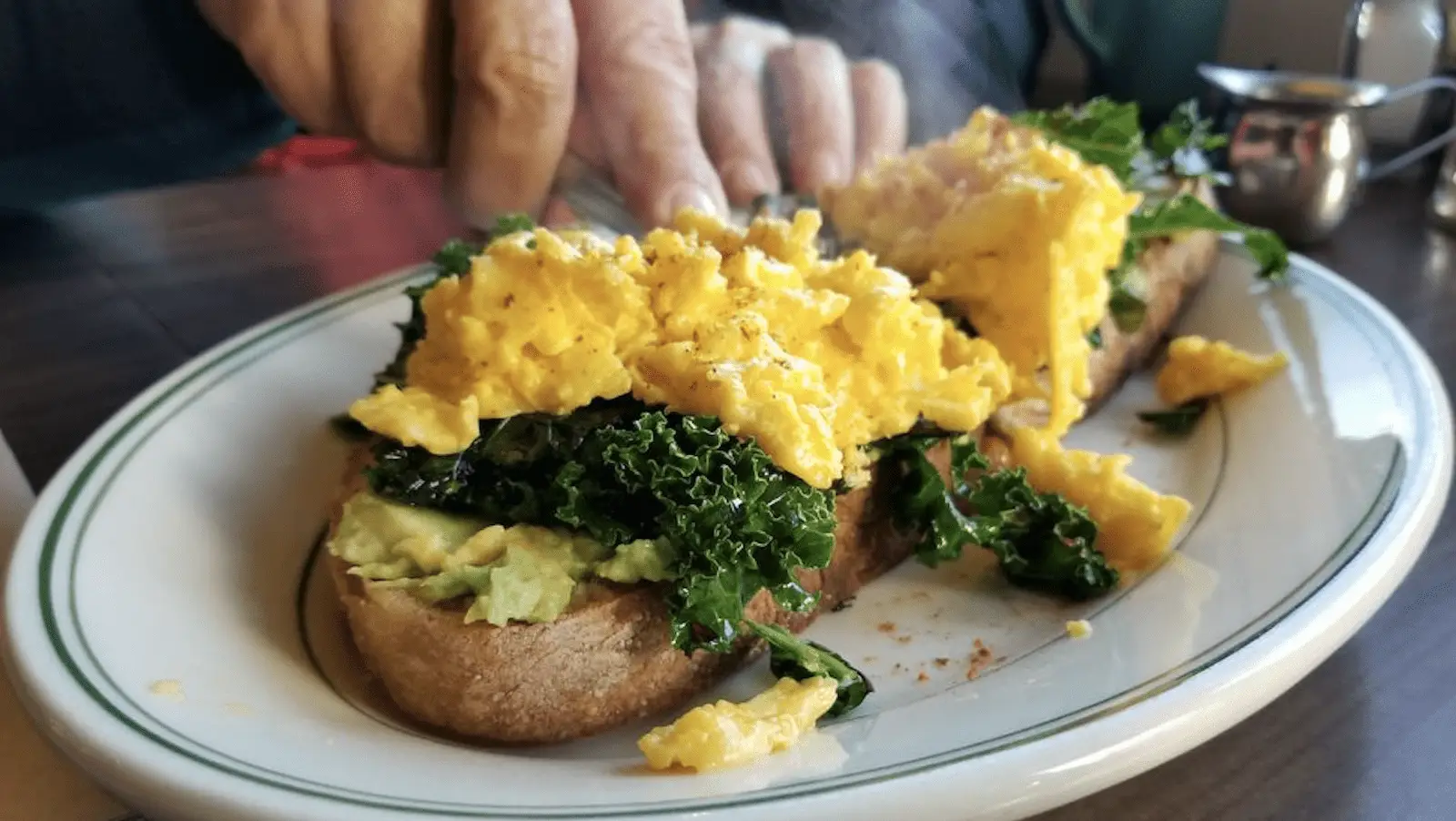 A close-up of a plate with toast topped with mashed avocado, sautéed kale, and scrambled eggs. A person's hands are visible in the background, reaching toward the food. The meal is on a white, oval plate with a green rim, set on a wooden table—truly the best breakfast North Bay has to offer.